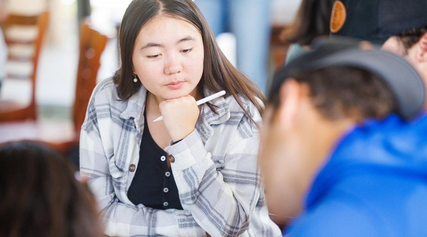 a student with long hair sits at a desk with a pencil in their hand